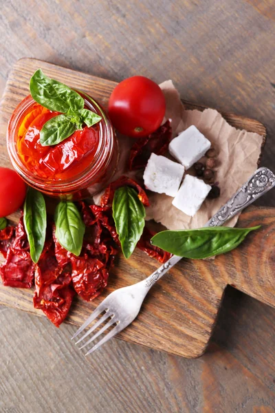 Tomates séchées au soleil dans un bocal en verre, feuilles de basilic et fromage feta sur planche à découper, sur fond en bois — Photo