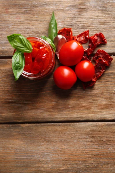 Sun dried tomatoes in glass jar, basil leaves on wooden background — Stock Photo, Image