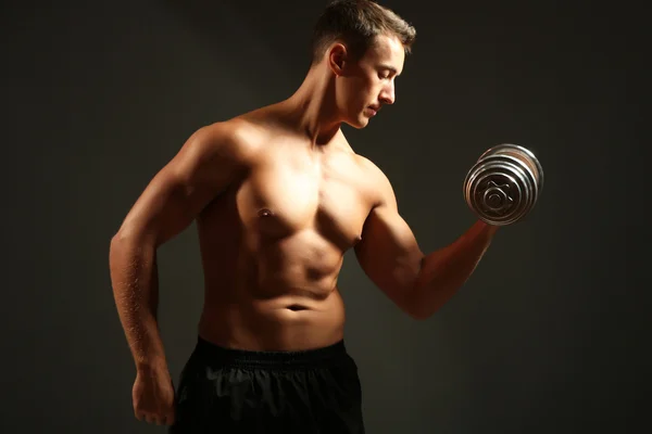 Handsome young muscular sportsman execute exercise with dumbbells on dark background — Stock Photo, Image