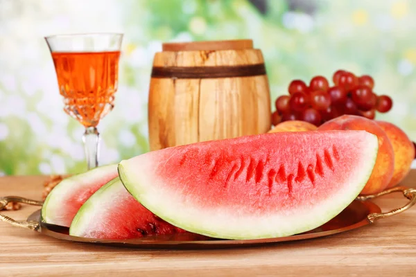 Composition of ripe watermelon, fruits, pink wine in glass and wooden barrel on  color wooden table, on bright background