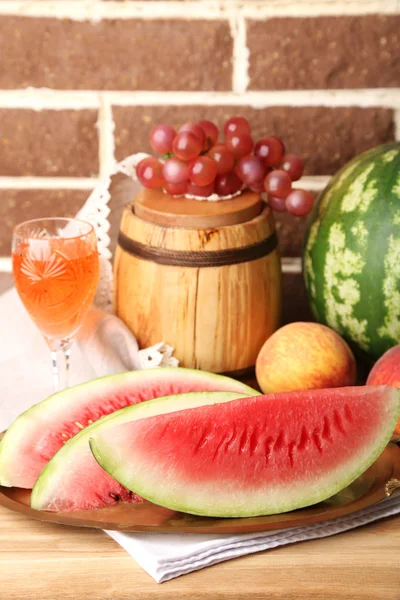 Sammansättningen av mogen vattenmelon, frukter, rosa vin i glas och trä fat på färg träbord, på tegelstenar bakgrund — Stockfoto