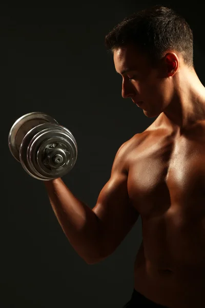 Handsome young muscular sportsman execute exercise with dumbbells on dark background — Stock Photo, Image