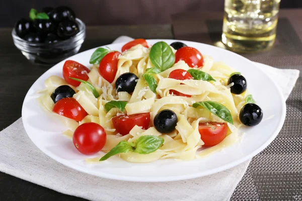 Spaghetti with tomatoes, olives and basil leaves on plate on tablecloth on wooden background — Stock Photo, Image