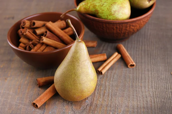 Ripe pears and cinnamon sticks in bowls, on wooden background — Stock Photo, Image