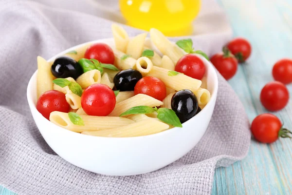 Pâtes avec tomates, olives, huile d'olive et feuilles de basilic dans un bol sur serviette sur fond en bois — Photo