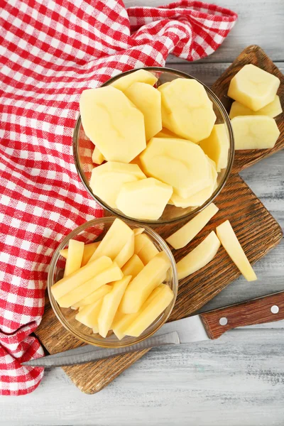 Patatas crudas peladas y en rodajas en cuencos de vidrio, sobre tabla de cortar, sobre fondo de madera de color — Foto de Stock