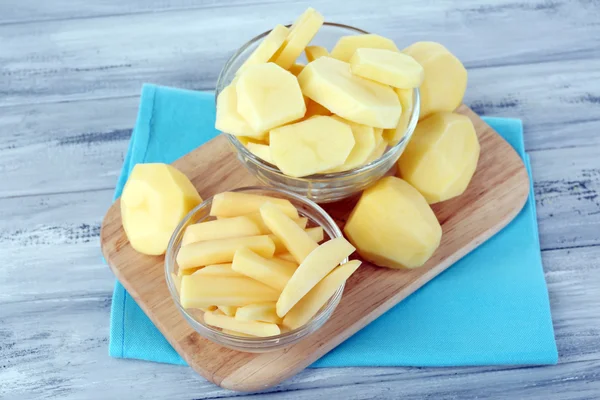 Rohe geschälte und in Scheiben geschnittene Kartoffeln in Glasschalen, auf Schneidebrett, auf farbigem Holzgrund — Stockfoto