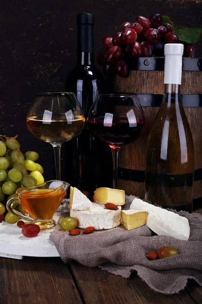 Cena compuesta de queso Camembert y Brie, miel, vino y uvas sobre tela de saco sobre soporte y barril de vino sobre mesa de madera sobre fondo marrón — Foto de Stock