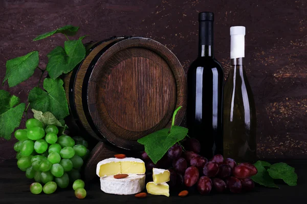 Wine in bottles and in goblet, Camembert and brie cheese, grapes and wooden barrel on wooden table on wooden background — Stock Photo, Image