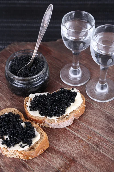 Fatias de pão com manteiga e caviar preto, jarra de vidro de caviar e par de pequenos copos em mesa de madeira no fundo escuro — Fotografia de Stock