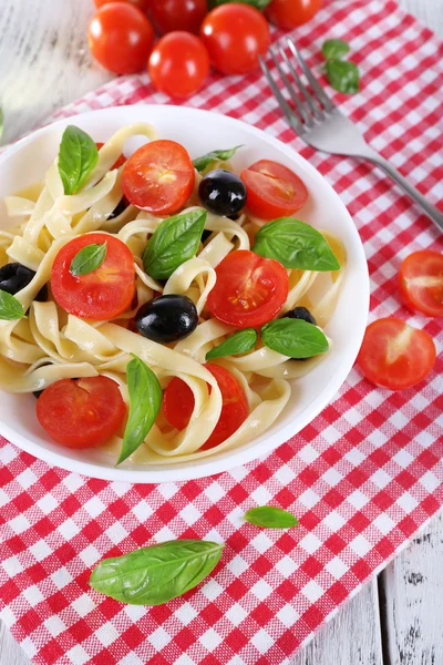 Спагетти с помидорами, оливками и листьями базилика на тарелке на салфетке на деревянном фоне — стоковое фото