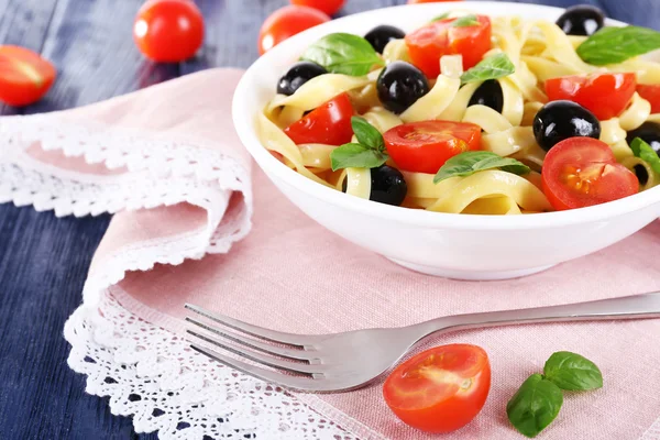 Спагетти с помидорами, оливками и листьями базилика на тарелке на розовой салфетке кружева на деревянном фоне — стоковое фото