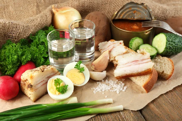 Bacon, fresh vegetables, boiled egg and bread on paper, glasses with vodka on wooden background. Village breakfast concept. — Stock Photo, Image