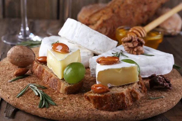 Camembert cheese on paper, grapes, nuts and honey in glass bowl on on cutting board on wooden background — Stock Photo, Image