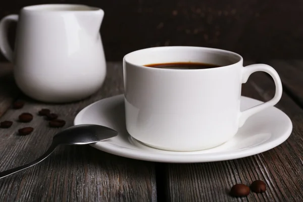 Cup of coffee with cream in milk jug and coffee beans on wooden table on dark background