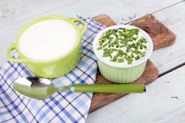 Green pan and plastic bowl of cream on a napkin on a cutting board on wooden background — Stock Photo, Image