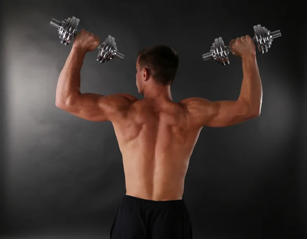 Handsome young muscular sportsman execute exercise with dumbbells on dark background — Stock Photo, Image