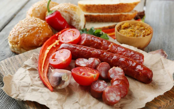 Smoked thin sausages and vegetables on cutting board, on wooden background — Stock Photo, Image