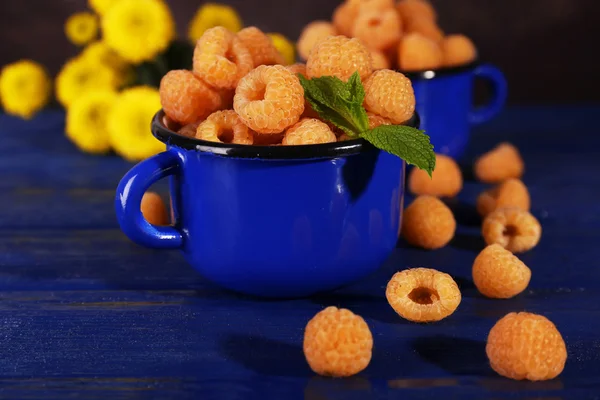 Yellow raspberries in dark blue mugs and wildflowers on wooden table on dark background — Stock Photo, Image