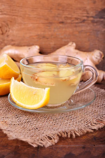 Cup of ginger drink and lemon on sackcloth napkin on wooden table on wooden background — Stock Photo, Image