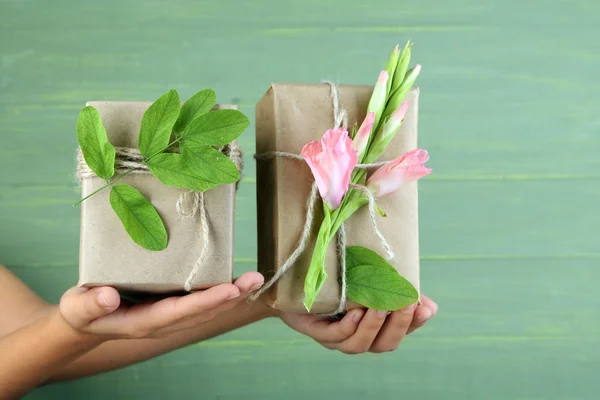 Mano femenina sosteniendo cajas de regalo artesanales de estilo natural con flores frescas y cordel rústico, sobre fondo de madera —  Fotos de Stock