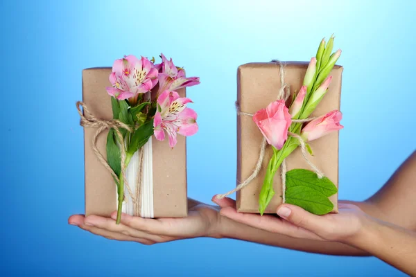 Mano femenina sosteniendo cajas de regalo artesanales de estilo natural con flores frescas y cordel rústico, sobre fondo de color —  Fotos de Stock