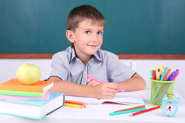 Schoolboy sitting in classroom on blackboard background — Stock Photo, Image