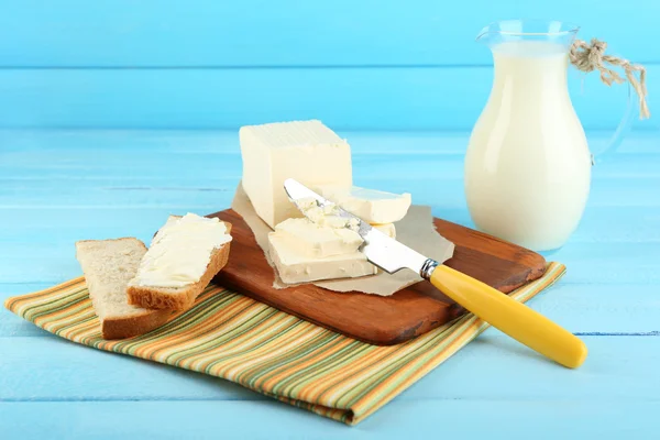 Fresh butter on cutting board and jug with milk, on color wooden background — Stock Photo, Image