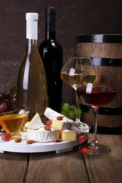 Večeře sestávající z hermelín sýr, med, víno a hrozny na stojan a vinný sud na dřevěný stůl na hnědé pozadí — Stock fotografie