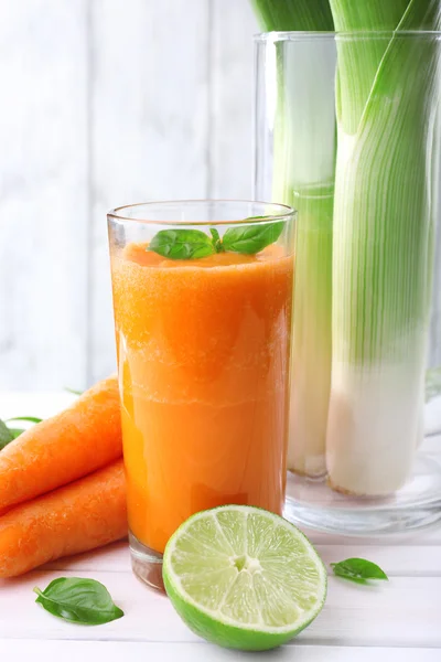 Склянка свіжого морквяного соку, весняна цибуля, лайм, трава і морква на столі перед дерев'яною стіною — стокове фото