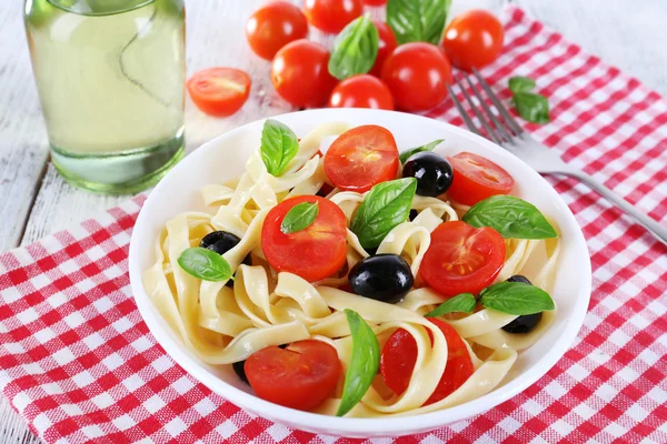 Spaghetti with tomatoes, olives and basil leaves on plate on napkin on wooden background — Stock Photo, Image