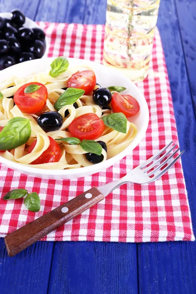 Spaghetti with tomatoes, olives and basil leaves on plate on napkin on wooden background — Stock Photo, Image