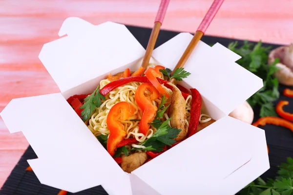 Chinese noodles and sticks in takeaway box on black mat on pink background — Stock Photo, Image