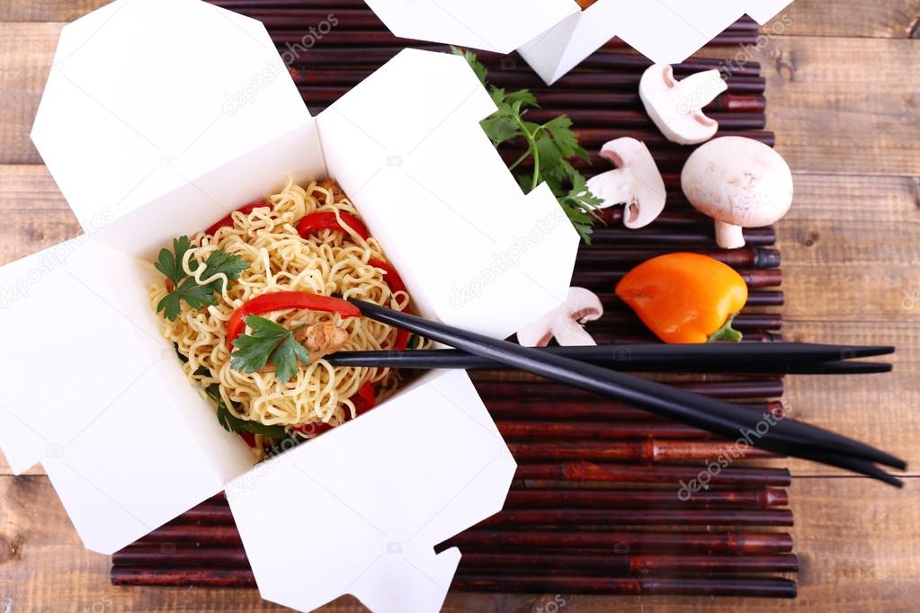 Chinese noodles in takeaway boxes on bamboo mat on wooden background