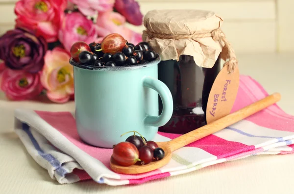 Rijpe zwarte bessen in mok en glazen pot met smakelijke jam op lichte houten achtergrond. — Stockfoto