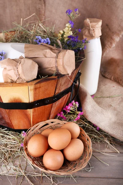 Big round basket with dried grass, milk and fresh eggs on sacking background — Stock Photo, Image