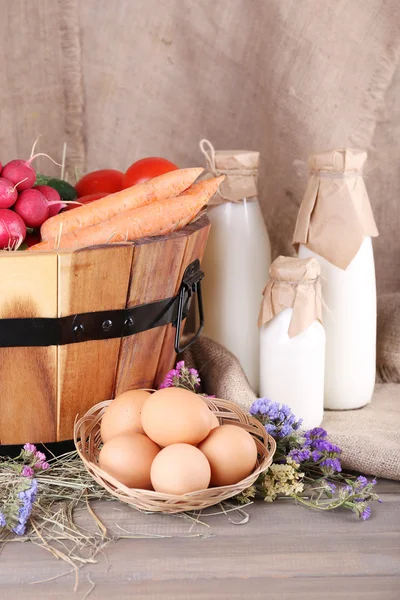 Big round basket with dried grass, vegetables, milk and fresh eggs on sacking background — Stock Photo, Image