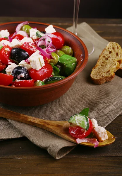 Greek salad served in brown bowl with slice of bread and glass of wine on napkin on wooden table on dark background — Stock Photo, Image