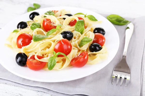 Spaghetti with tomatoes, olives and basil leaves on plate on napkin on wooden table — Stock Photo, Image