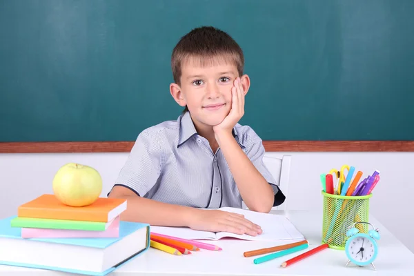 Schoolboy sitting in classroom on blackboard background — Stock Photo, Image