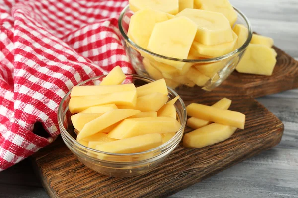 Rohe geschälte und in Scheiben geschnittene Kartoffeln in Glasschalen, auf Schneidebrett, auf farbigem Holzgrund — Stockfoto