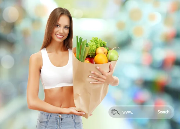 Shopping concept. Beautiful young woman with vegetables and fruits in shopping bag on shop background — Stock Photo, Image