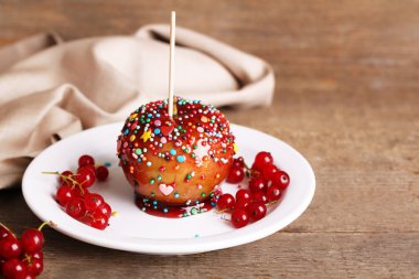 Sweet caramel apple on stick with berries, on wooden table clipart