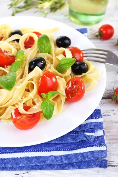 Spaghetti with tomatoes, olives and basil leaves on plate on napkin on wooden background — Stock Photo, Image