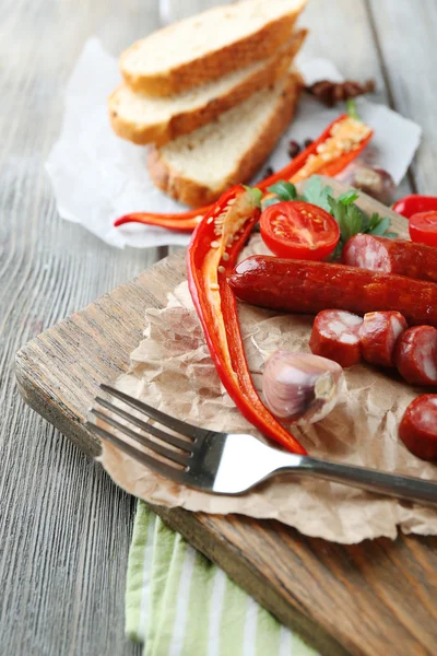 Smoked thin sausages and vegetables on cutting board, on wooden background — Stock Photo, Image