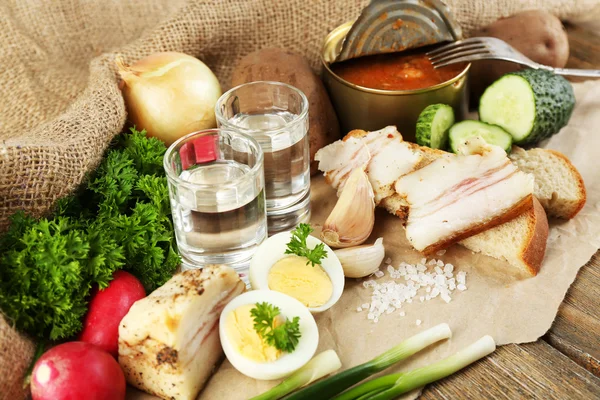 Bacon, fresh vegetables, boiled egg and bread on paper, glasses with vodka on wooden background. Village breakfast concept. — Stock Photo, Image