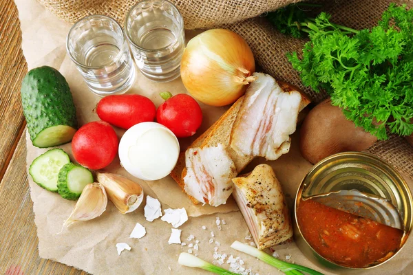 Bacon, fresh vegetables, boiled egg and bread on paper, glasses with vodka on wooden background. Village breakfast concept. — Stock Photo, Image