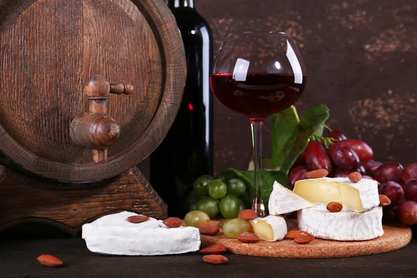 Kveldsmat bestående av kamemberost, vin og druer på nedskjæringstavle og vintønne på trebord på brun bakgrunn – stockfoto