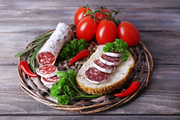 French salami with tomatoes, bread and parsley on wicker mat on wooden background — Stock Photo, Image