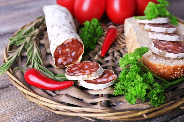 French salami with tomatoes, bread and parsley on wicker mat on wooden background — Stock Photo, Image